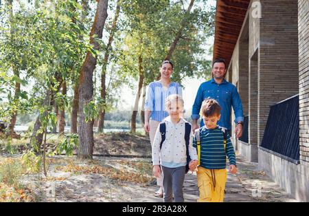 Junge Eltern sehen ihre süßen Kinder zur Schule gehen. Lächelnde eltern, Eltern, Tochter und Sohn gehen die Treppen auf der Terrasse ihres Hauses hinunter. Junge und Mädchen i Stockfoto