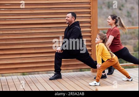 Glückliche Familien dehnen sich draußen aus. Attraktive mutter, attraktiver Vater und süße Tochter, die Ausfallschritte macht. Das junge Paar und sein Kind trainieren im Park Stockfoto