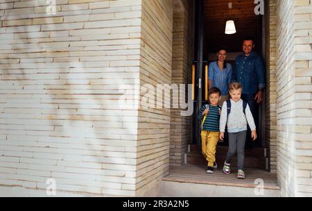 Junge Eltern sehen ihre süßen Kinder zur Schule gehen. Lächelnde eltern, Eltern, Tochter und Sohn gehen die Treppen auf der Terrasse ihres Hauses hinunter. Junge und Mädchen i Stockfoto