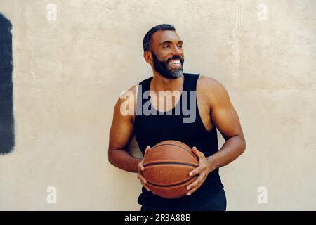 Ein bärtiger Sportler trainiert drinnen und dribbelt einen Basketball. Er trägt ein ärmelloses Tanktop und sieht auf seinem Streben nach Erfolg entschlossen aus. Stockfoto