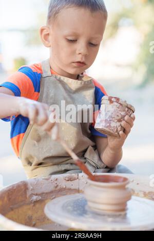 Hände eines jungen Töpfers, der ein Erdenglas am Kreis kreiert, Nahaufnahme. Der kleine lächelnde Junge wurde produziert Stockfoto