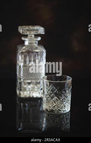 Leeres Glas für Whiskey oder Bourbon mit einer quadratischen Karaffe aus Kristall auf der schwarzen reflektierenden Oberfläche Stockfoto