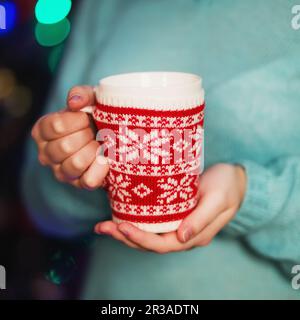 Ein Becher mit Strickzeug in weiblichen Händen. Das Mädchen in einem gemütlichen Strickpullover trinkt C. Stockfoto