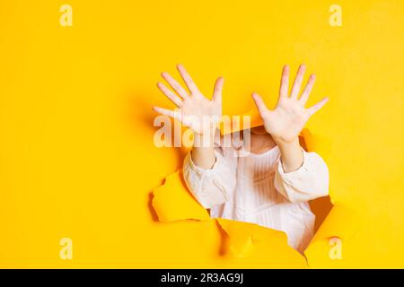 Kinderhände zeigen zehn Finger durch ein eingerissenes Loch in gelbem Papier mit Kopierraum. Stockfoto