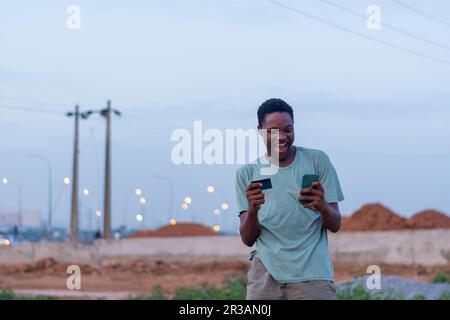 Glückliche afrikanische Jugend, die sein Handy und seine Kreditkarte benutzt, um online zu bezahlen. Kauf- und Einkaufskonzept Stockfoto