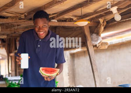 Afrikanischer Händler mit Wassermelone auf einem lokalen afrikanischen Markt und einem Mobiltelefon mit einem leeren weißen Bildschirm, für Werbeschriften, Werbung Stockfoto