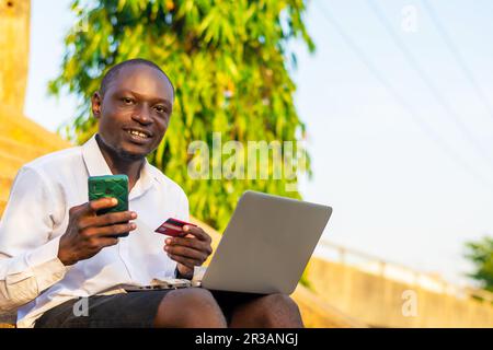 Kopieren Sie das Weltraumbild eines jungen, gutaussehenden Geschäftsmanns, der ein Notebook auf dem Schoß trägt und sein Smartphone und seine Kreditkarte für Transaktionen verwendet Stockfoto