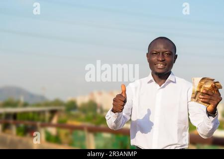 Ein glücklicher afrikaner, der mit einem strahlenden Lächeln steht und Bargeld hält, während er direkt in die Kamera schaut, Daumen nach oben zeigt und Zeichen der Anerkennung zeigt. Stockfoto