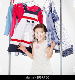 Kleine Frau probiert neue Kleidung auf weißem Hintergrund. Ein kleines Mädchen wählt ein Kleid. Kleines Mädchen und Gerinnsel Stockfoto