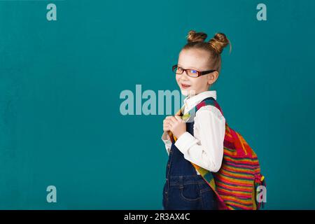 Lustiges kleines Mädchen mit einem großen Rucksack, das sich an der türkisfarbenen Wand amüsiert. Schulkonzept. B Stockfoto