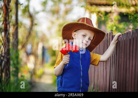 Porträt eines kleinen Jungen, der in einem großen Hut steht, mit einem Strauß Tulpen am Holzzaun im C. Stockfoto