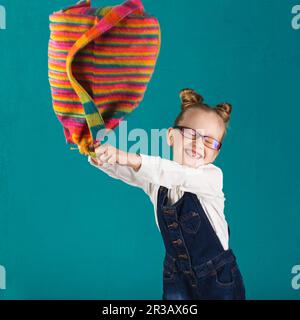 Lustiges kleines Mädchen mit einem großen Rucksack, das sich an der türkisfarbenen Wand amüsiert. Schulkonzept. B Stockfoto