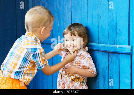 Bruder kitzelt ihre Schwester. Kleine Kinder spielen gegen blaue Holzwand Stockfoto
