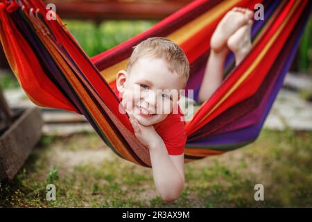 Schöner fröhlicher kleiner Junge, der in einer Hängematte ruht und Spaß hat. Positive Emotionen. Stockfoto