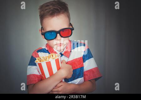 Ein kleiner Junge sieht sich einen Film in 3D-Brillen im Kino oder zu Hause an. Das kleine Kind isst Popcorn über grauem Backen Stockfoto