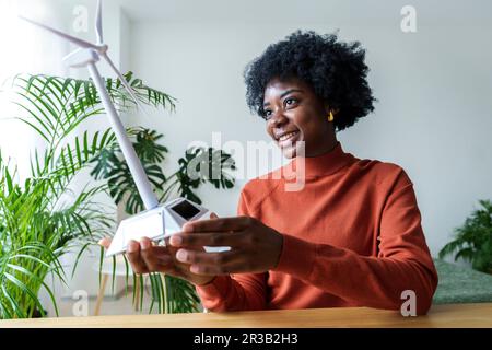Glückliche Geschäftsfrau mit Windturbinenmodell, die am Schreibtisch sitzt Stockfoto