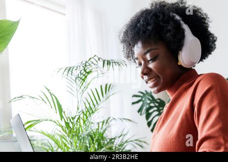 Glückliche junge Geschäftsfrau mit kabellosen Kopfhörern, die auf einem Laptop arbeitet Stockfoto