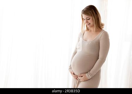 Lächelnde schwangere Frau, die zu Hause neben dem Vorhang steht Stockfoto
