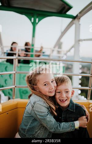 Lächelndes Mädchen, das den Bruder umarmt, der im Riesenrad im Vergnügungspark sitzt Stockfoto