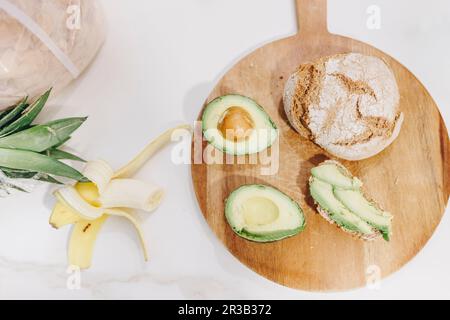 Halb gefressene Banane mit Avocado und Brot auf dem Schneidebrett Stockfoto