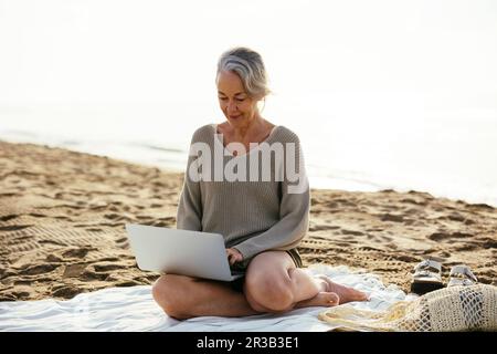 Erwachsene Frau, die am Strand mit einem Laptop sitzt Stockfoto