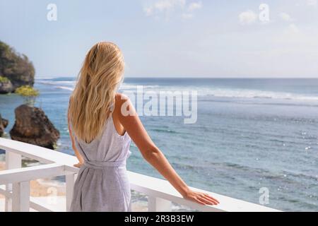 Eine blonde Frau, die das Meer ansieht und neben dem Geländer steht Stockfoto