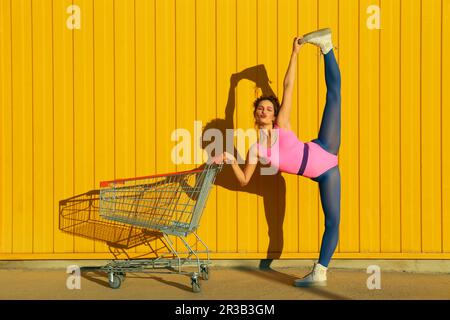 Balletttänzerin, die vertikal am Einkaufswagen vor der gelben Wand spaltet Stockfoto
