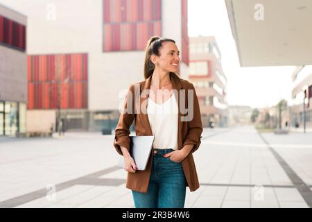Lächelnde Geschäftsfrau, die ihren Laptop in der Hand hält und auf dem Fußweg läuft Stockfoto