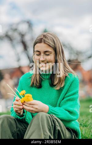 Lächelndes Teenager-Mädchen, das einen Kranz mit gelben Löwenzahnen auf dem Gras macht Stockfoto