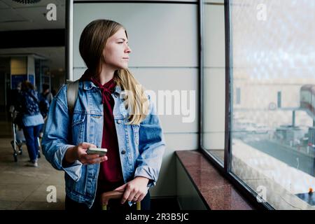 Junge Frau mit Smartphone, die aus dem Fenster am Flughafen schaut Stockfoto