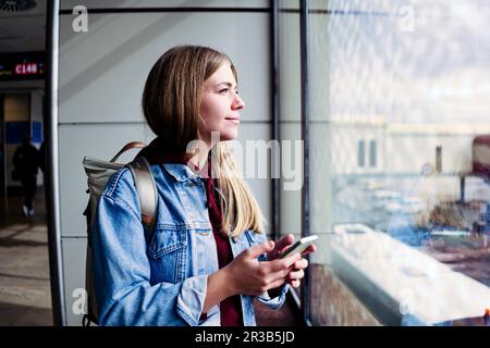 Junge Frau, die aus dem Fenster schaut und ein Smartphone am Flughafen hält Stockfoto