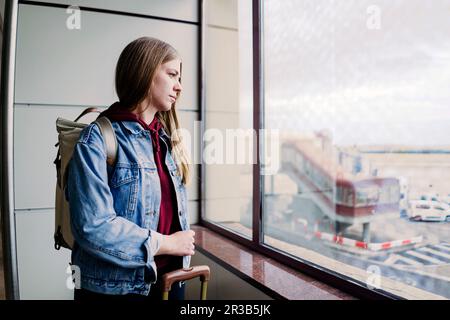 Junge Frau mit Rucksack, die aus dem Flughafenfenster schaut Stockfoto