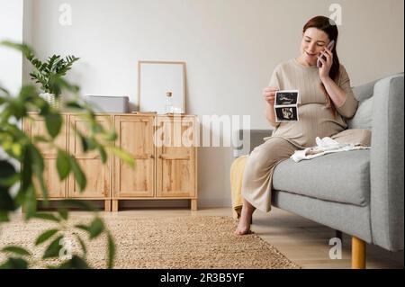 Schwangere Frau, die mit einem Smartphone spricht und Ultraschallbilder zu Hause hält Stockfoto