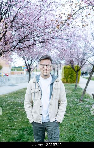 Ein lächelnder Mann stand mit Händen in Taschen neben dem Kirschblütenbaum im Park Stockfoto