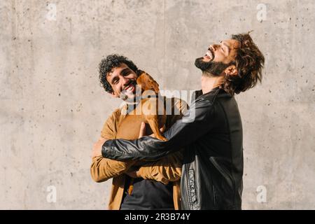 Glückliches schwules Paar, das seine Freizeit mit dem Hund vor der Wand verbringt Stockfoto