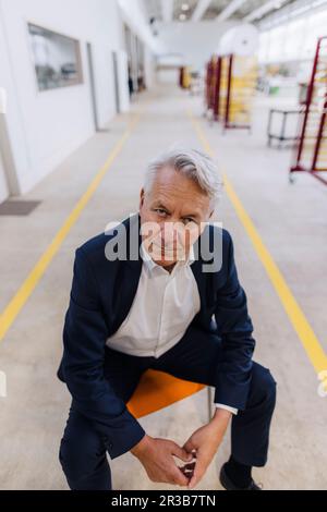 Ein seriöser Geschäftsmann, der in der Fabrik auf einem Stuhl sitzt Stockfoto