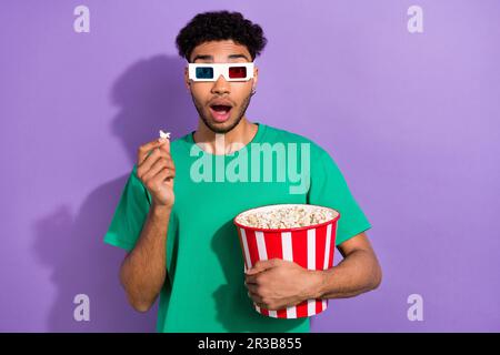 Foto eines beeindruckten, schockierten Mannes, der ein grünes T-Shirt trägt und Popcorn isst und sich einen Film mit violettem Hintergrund ansieht Stockfoto