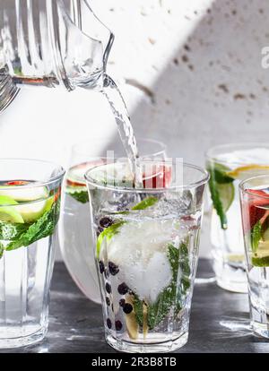 Verschiedene Gläser Wasser gefüllt mit Obst, Minze und Ingwer, wobei Wasser aus einem Krug in ein Glas gegossen wird Stockfoto