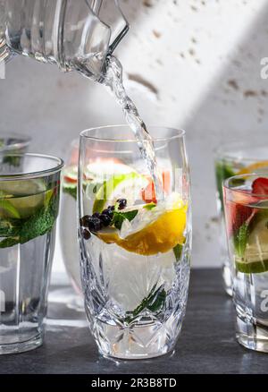 Verschiedene Gläser Wasser gefüllt mit Obst, Minze und Ingwer, wobei Wasser aus einem Krug in ein Glas gegossen wird Stockfoto