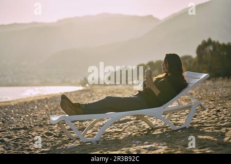 Frau, die sich auf dem Liegestuhl zurücklehnt und Thermoskanne am Strand hält Stockfoto