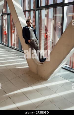 Geschäftsmann, der Sprachnachrichten über ein Smartphone sendet und sich auf eine V-förmige Säule im Gebäude lehnt Stockfoto