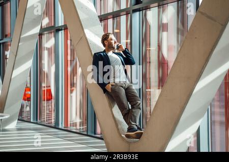 Ein Geschäftsmann, der am Telefon telefoniert und sich auf eine V-förmige Säule im Gebäude lehnt Stockfoto