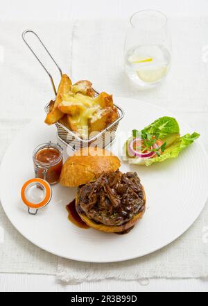 Schweinefleisch in einem weißen Brötchen mit Barbecue-Sauce, serviert mit Chips mit geschmolzenem Käse, einem Topf Tomatenrelish und einem grünen Salat Stockfoto