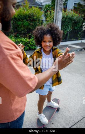 Ein Mädchen, das auf dem Skateboard steht und sich an den Händen seines Vaters hält Stockfoto