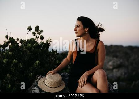 Lächelnde Frau mit Hut, die auf einem Stein sitzt Stockfoto