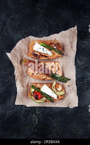 Hausgemachtes getoastetes Brot mit grünem Spargel, eingewickelt in Speck, Käse, Avocado-Scheiben, gegrillter Tomatenkirsche und Nüssen Stockfoto