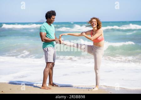 Ein Mann, der eine Freundin beim Stretchen und Stehen auf einem Bein an der Küste unterstützt Stockfoto