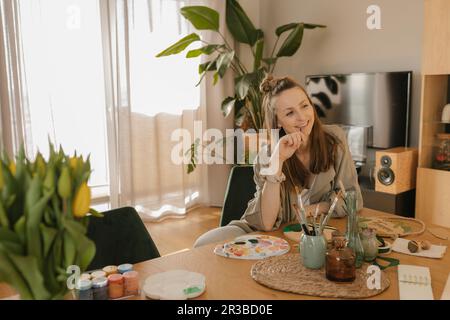 Lächelnde, nachdenkliche junge Frau, die am Tisch malt Stockfoto