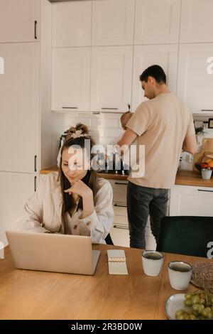 Frau, die in der Küche an einem Notebook arbeitet, mit Familie im Hintergrund zu Hause Stockfoto