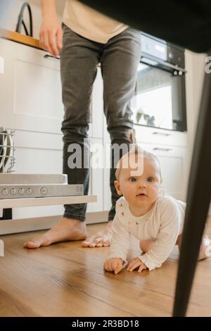 Süßes Mädchen, das auf dem Boden krabbelt, von Vater, der zu Hause Utensilien in die Spülmaschine lädt Stockfoto